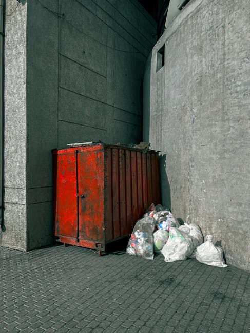 An outdoor urban scene showing a large, rectangular metal rubbish skip with a weathered, red finish positioned against a dark green tiled wall and a light grey concrete wall. The surface of the skip appears worn with visible patches of rust and peeling paint. Surrounding the base of the skip are several small to medium-sized plastic rubbish bags filled with waste, predominantly white and transparent, with some showing green and black items inside. The bags are loosely gathered and placed on a paved surface composed of grey rectangular bricks arranged in a herringbone pattern. The environment suggests a back alley or side street location, with minimal natural light and no visible signage. The overall setting indicates a private waste collection area where waste clearance services such as waste clearance Watford may be involved in removing accumulated rubbish from commercial or residential sites to support local rubbish removal needs.