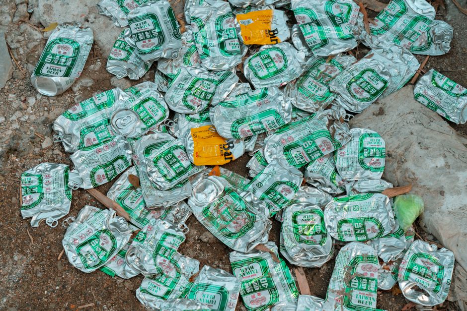 A collection of crushed aluminum cans scattered across a dirt ground surface outdoors. The cans are silver with green and white labels, indicating they are Taiwanese beer cans, and are crumpled and stacked irregularly. Some cans are partially flattened, revealing characteristic ridges and pull-tab tops. Among the cans, there are small pieces of debris and organic matter, including dried leaves and fragments of wood, enhancing the cluttered appearance. A yellow and orange paper label is visible attached to one of the cans. The background shows a mix of loose soil and scattered natural debris, suggesting an untidy outdoor area where waste has accumulated. The scene reflects a typical outdoor environment where beverage cans have been discarded or left, suitable for waste clearance or rubbish removal services focusing on container waste cleanup in outdoor settings.