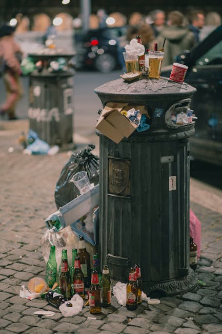 An outdoor urban scene showing a large, rectangular metal rubbish skip with a weathered, red finish positioned against a dark green tiled wall and a light grey concrete wall. The surface of the skip appears worn with visible patches of rust and peeling paint. Surrounding the base of the skip are several small to medium-sized plastic rubbish bags filled with waste, predominantly white and transparent, with some showing green and black items inside. The bags are loosely gathered and placed on a paved surface composed of grey rectangular bricks arranged in a herringbone pattern. The environment suggests a back alley or side street location, with minimal natural light and no visible signage. The overall setting indicates a private waste collection area where waste clearance services such as waste clearance Watford may be involved in removing accumulated rubbish from commercial or residential sites to support local rubbish removal needs.