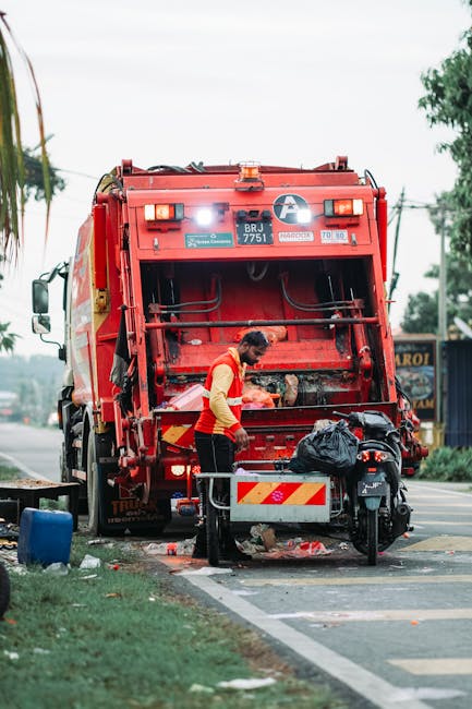 A worker wearing a high-visibility yellow and red uniform is standing at the rear of a large red waste collection vehicle, engaged in rubbish clearance on a street. The vehicle, likely a specialist waste truck, features a metal hopper with a textured, painted surface, and various reflective hazard markings in yellow and red. The worker appears to be loading or managing discarded items, with a black trash bag positioned on or near the vehicle's loading area. In the foreground, there is a small, partly opened metal waste container or trolley, and some scattered debris on the pavement. The scene is set outdoors in an urban or semi-urban environment, with a background that includes trees, utility poles, and a building with signage, under an overcast sky. The scene reflects an independent waste removal process, possibly offered by Waste Clearance Watford, involving manual handling and on-site disposal of rubbish.