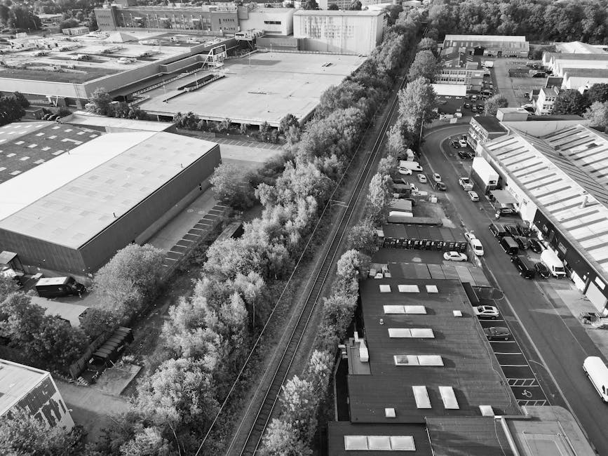 An aerial view of an urban area showing a railway track running through the center of the image, flanked by a line of trees with dense foliage on both sides. To the left of the railway, there is a large industrial or warehouse building with a flat, light-colored roof and surrounding parking areas, some with vehicles parked along the perimeter. To the right, there are commercial or office buildings with multiple flat roofs and a parking lot filled with various cars and vans. In the background, additional industrial buildings and a mix of trees and smaller structures are visible, with the environment appearing organized and well-maintained. The overall scene highlights a typical cityscape with transportation infrastructure and nearby commercial properties, consistent with areas where private waste management services, like those offered by Waste Clearance Watford, might operate, especially for on-site clearance and rubbish removal in urban environments.